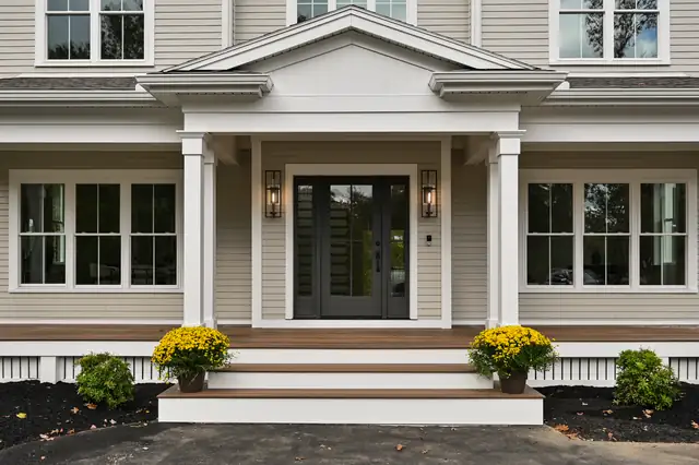 Front view of a two-story new construction home with greige siding, white columns, a black front door and fall mums on the steps