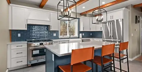 Ipswich, MA kitchen with a navy island, orange counter stools, wood ceiling beams, blue tile backsplash and white cabinets